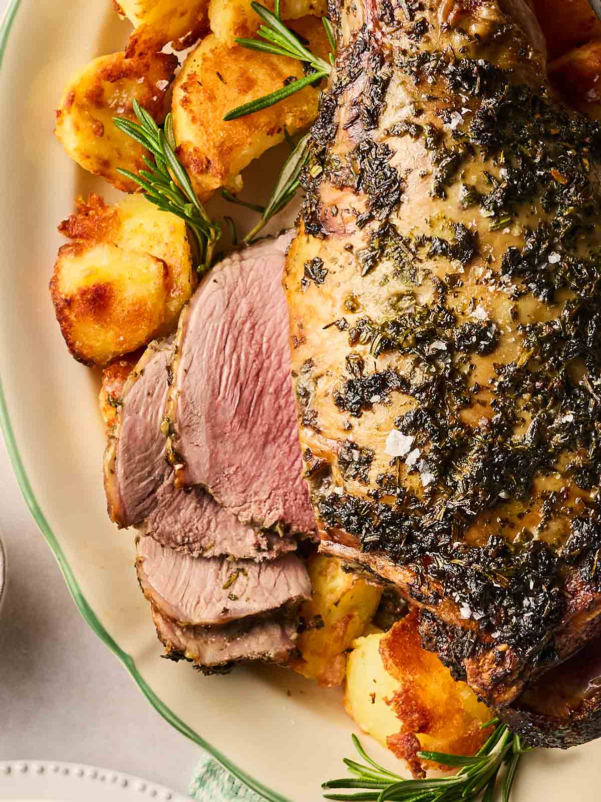 A close up of a half sliced roasted lamb leg with a green herb rub on top, with roast potatoes, on a platter.