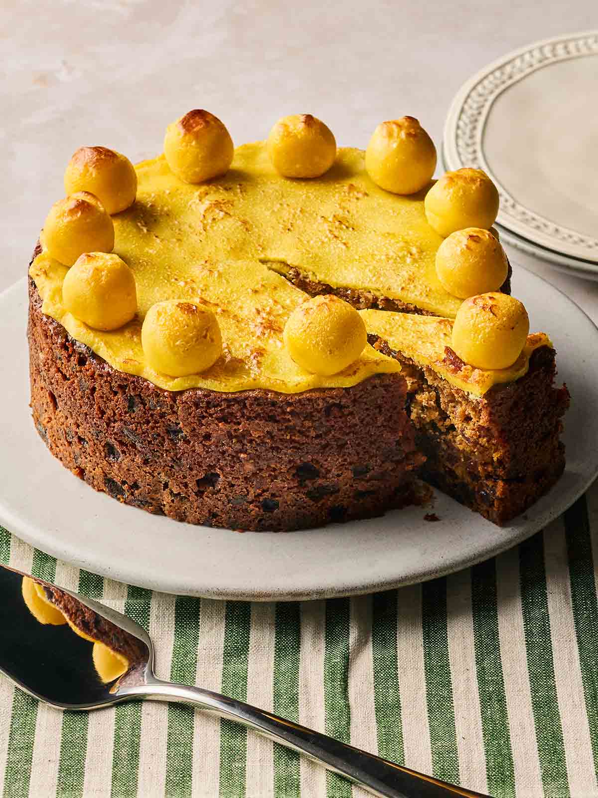 A traditional Simnel Cake on a white plate, with one slice cut out, ready to serve.