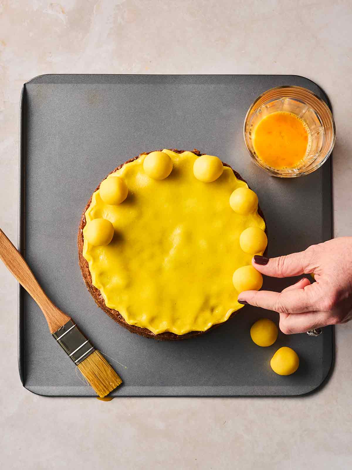 A Simnel Cake on a baking tray, being decorated with marzipan balls with one hand.