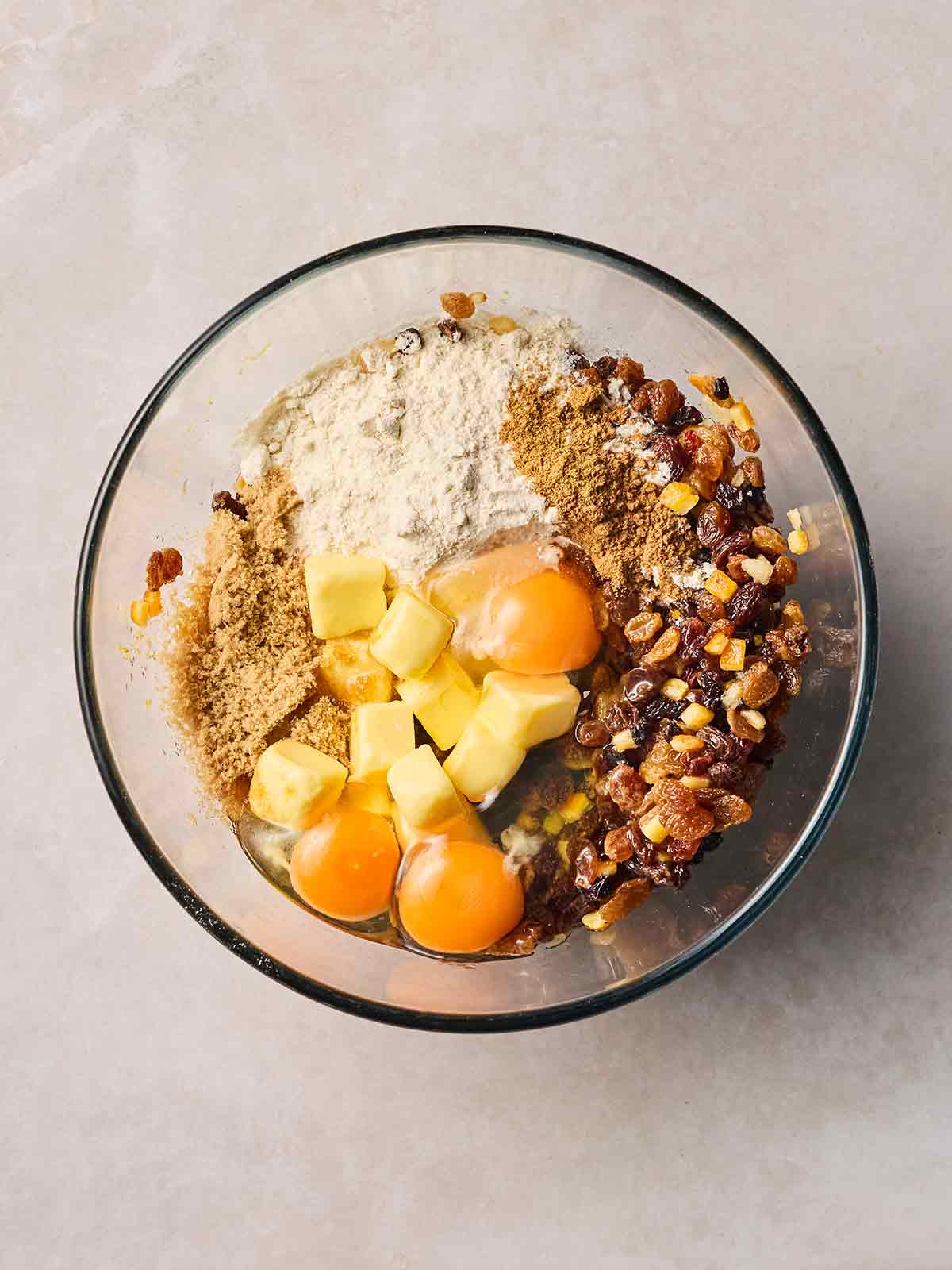 A glass bowl with lots of cake ingredients, not yet mixed, including flour, sugar, eggs, mixed fruit and baking powder.