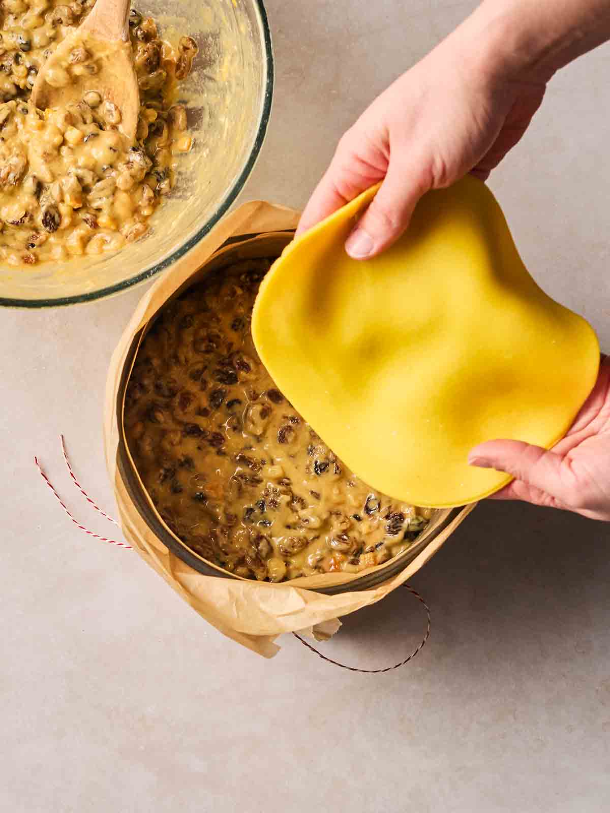 Half a mixture of fruit cake batter in a lined tin, with the other half still in the bowl, and two hands about to place a round of marzipan over the cake in the tin.