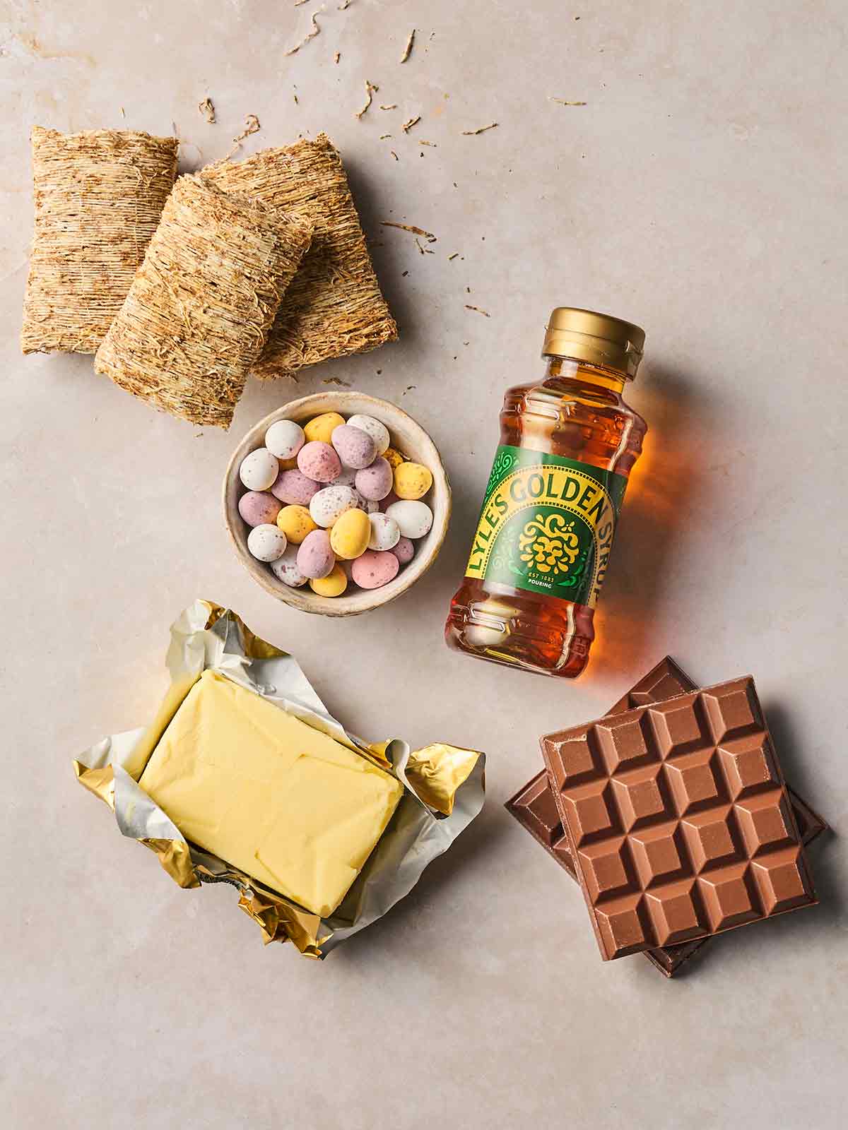 The ingredients for making chocolatey Easter Nests laid out on a white counter, including shredded wheat, golden syrup, mini eggs, butter and chocolate.