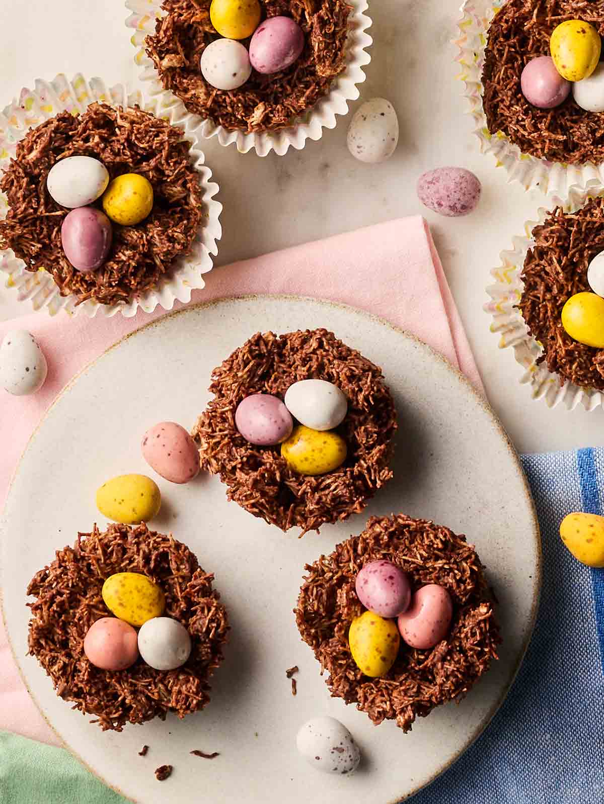 A batch of chocolate Easter Nests with mini eggs on top on a table, with scattered mini eggs.