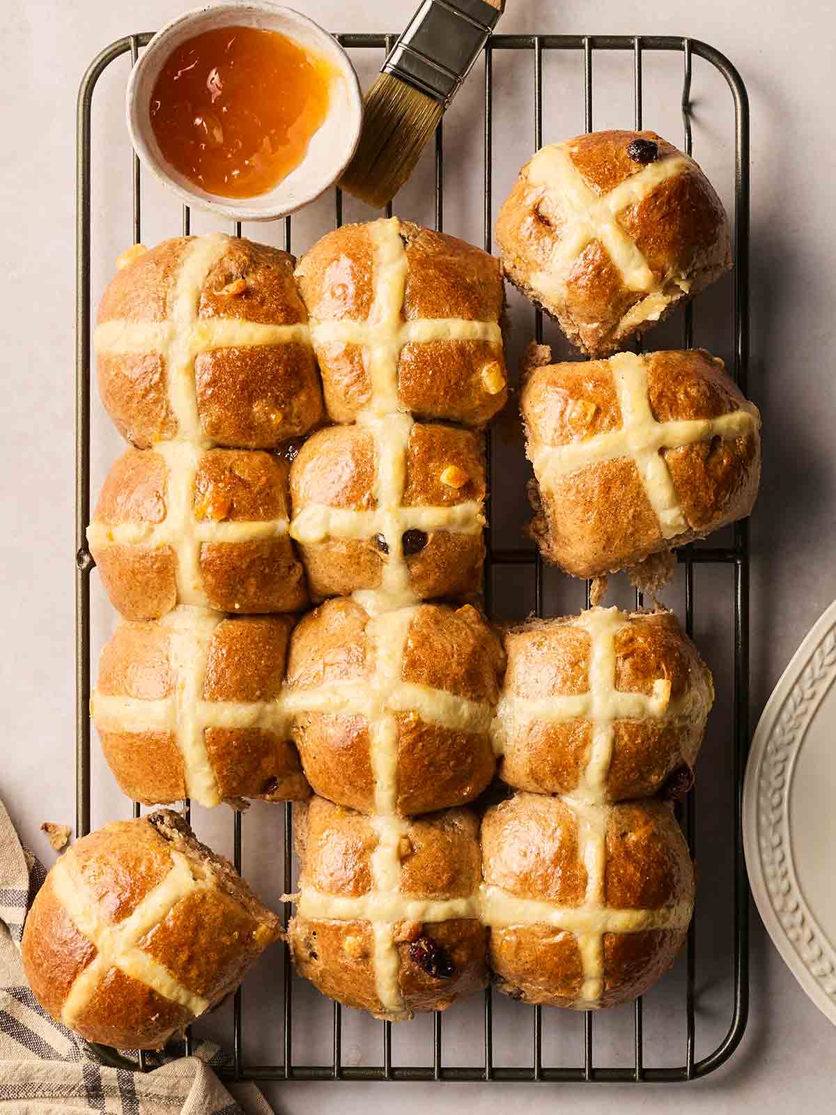 A wire rack with baked homemade Hot Cross buns on, with a bowl of apricot jam and a brush to add at the end.