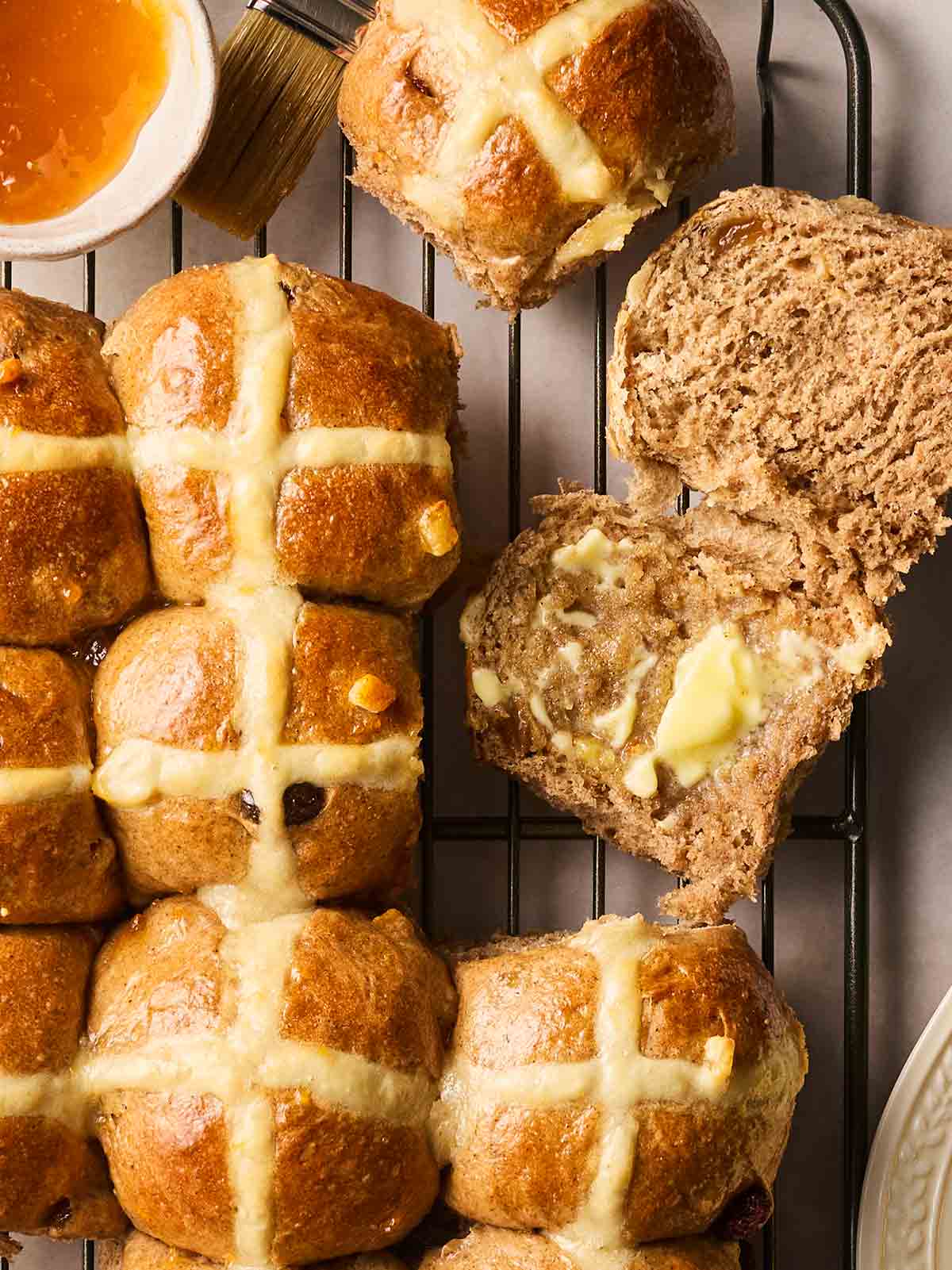 A close up of rows of just-baked Hot Cross Buns, with one sliced in half with butter spread on, ready to eat.