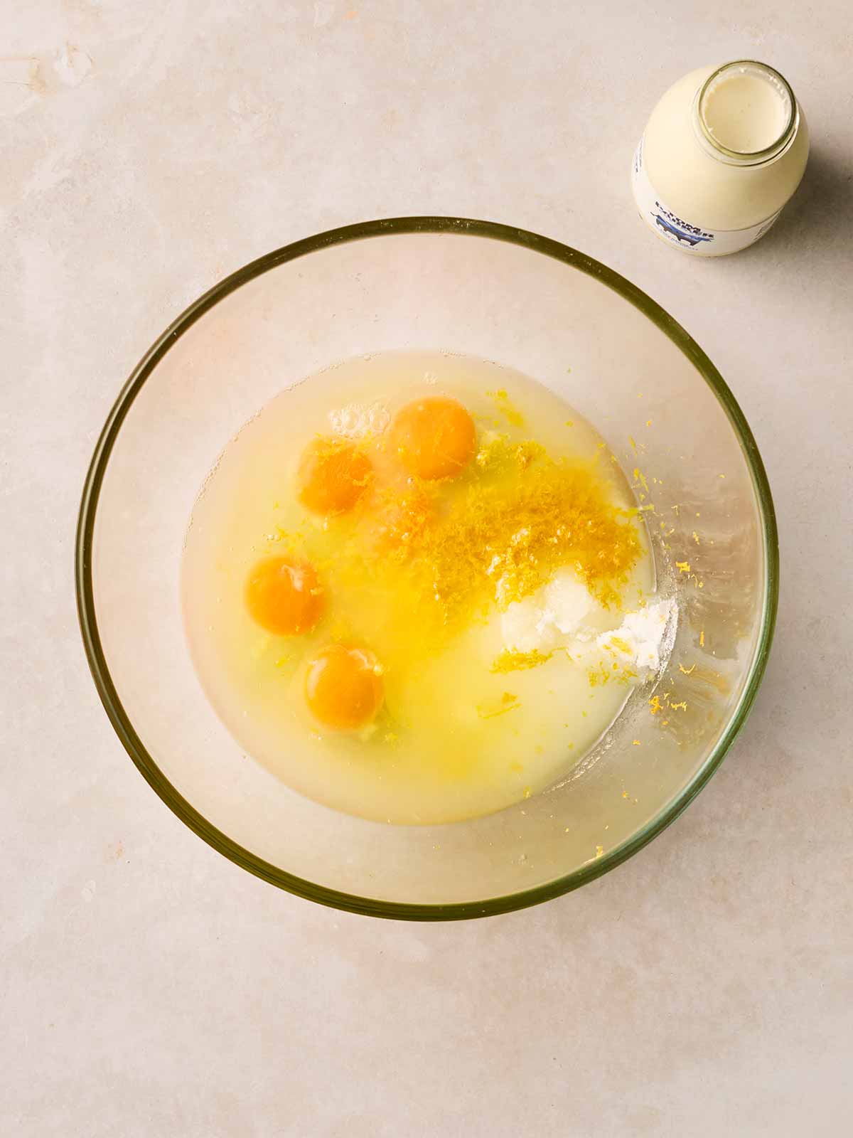 A glass bowl with cream, four cracked eggs and caster sugar in side, ready to be mixed.