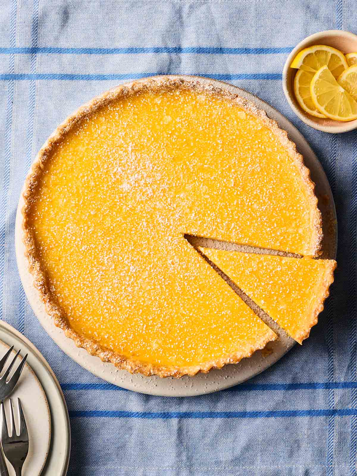 A classic lemon tart on a plate, on a blue tablecloth, with a slice cut out of it.