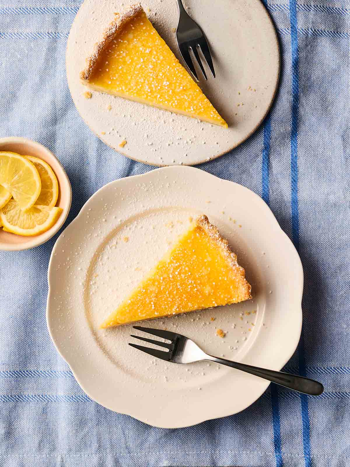 Two slices of classic lemon tart with shortcrust pastry on white plates with dessert forks, all on a blue tablecloth.