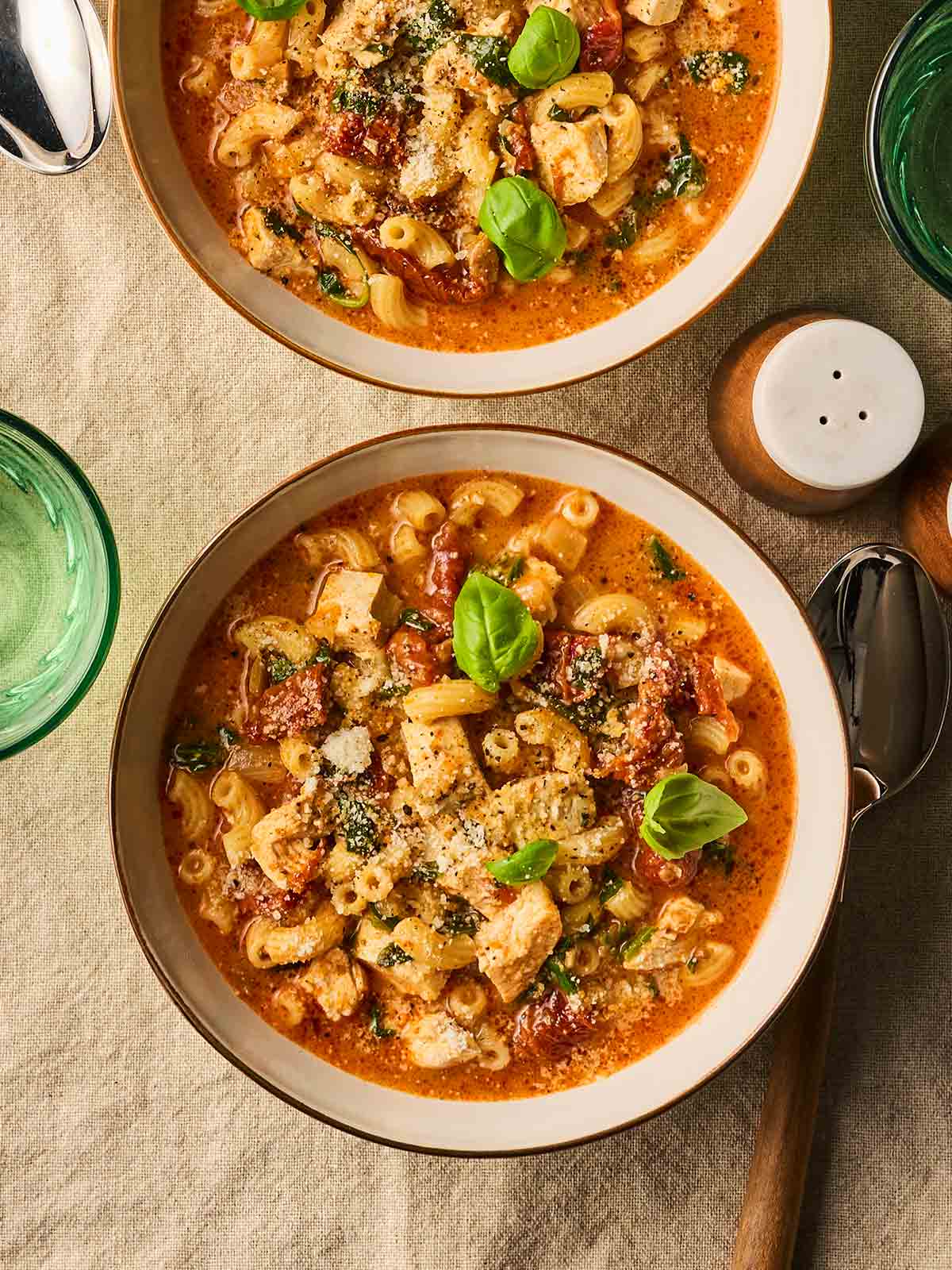Two bowls of soup on a table, with chicken, spinach, pasta and a tomatoey sauce.