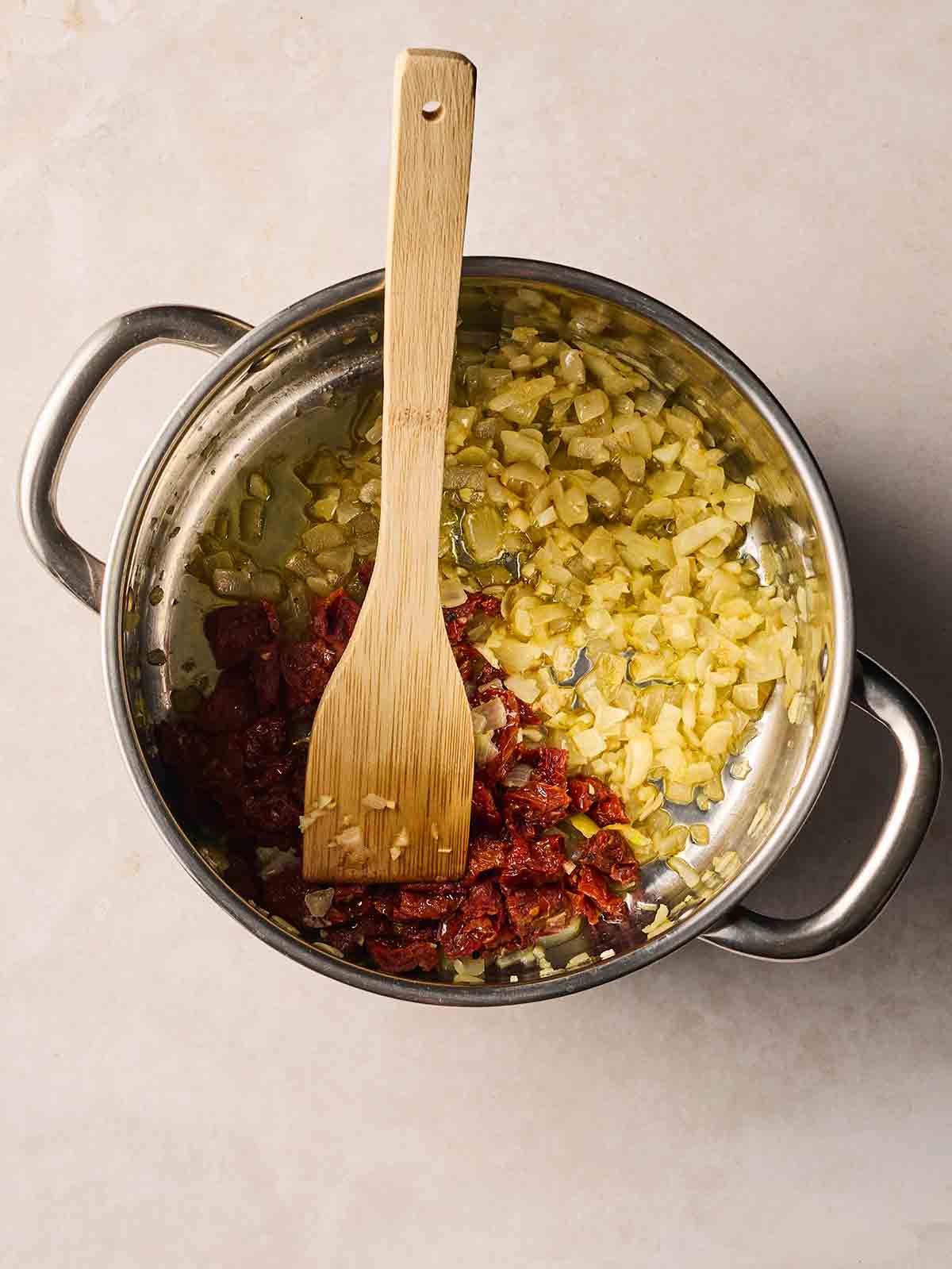 A silver pan with frying onions in oil and sun-dried tomatoes, with a wooden spoon to stir inside.