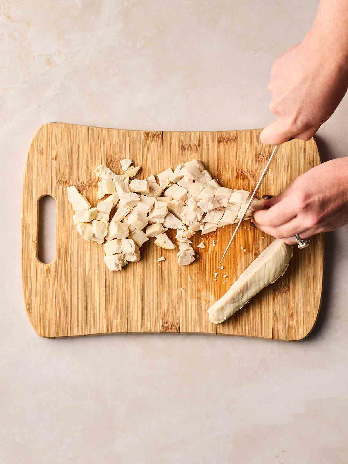 Two hands chopping up a cooked chicken breast on a wooden board with a knife into bite-sized chunks.
