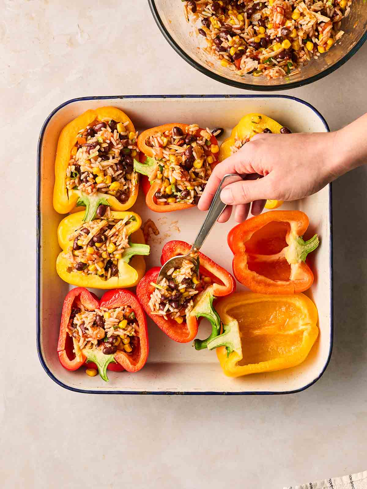 A hand spooning a rice mixture into halved peppers in a roasting dish.