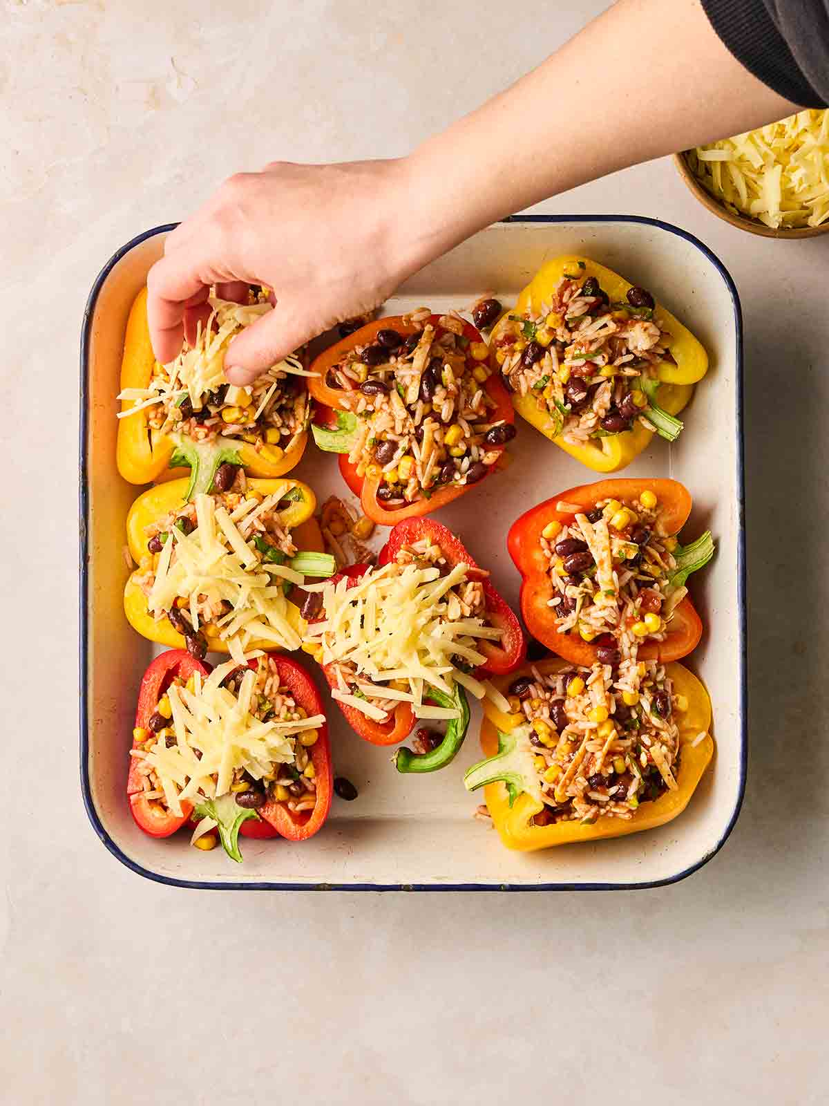 An oven dish filled with stuffed peppers, with a hand adding grated cheese on top for the final recipe step.