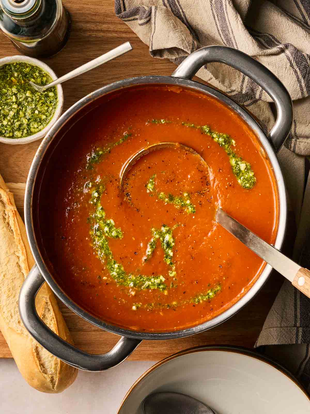A big pan of rich tomato soup with pesto swirled through, with a ladle in and on top of a table, ready to serve, with a baguette at the side.