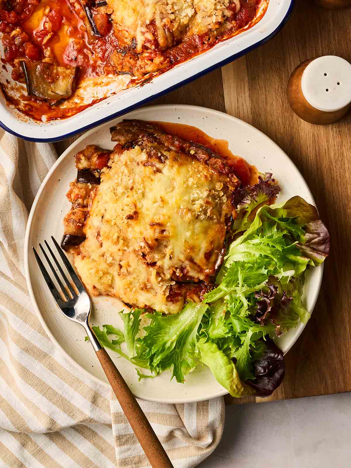 A portion of homemade Aubergine Parmigiana on a plate with salad and a fork, with the rest of the dish to the side on a table, ready to serve.
