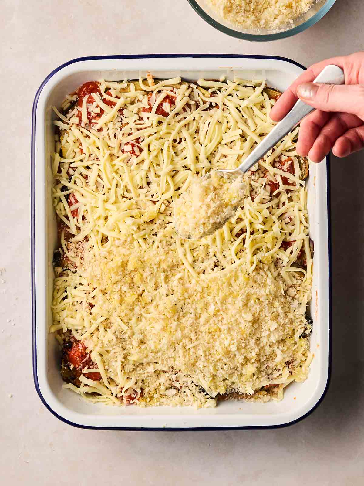 A hand sprinkling breadcrumbs with a spoon over an oven dish of unbaked Aubergine Parmigiana.