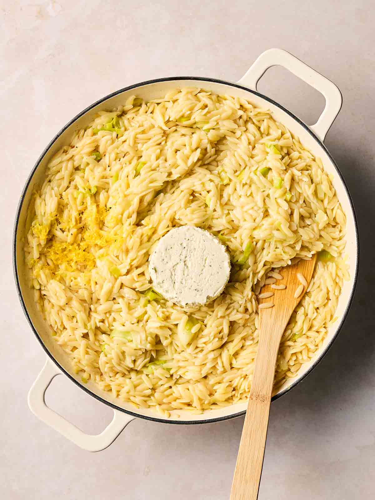 A pan of cooked orzo and leeks, with a circle of Boursin in the middle, ready to be stirred with a wooden spoon.