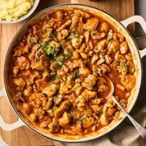 A large pan filled with Chicken Chasseur, with a spoon in ready to serve, on a table with mashed potato to the side.