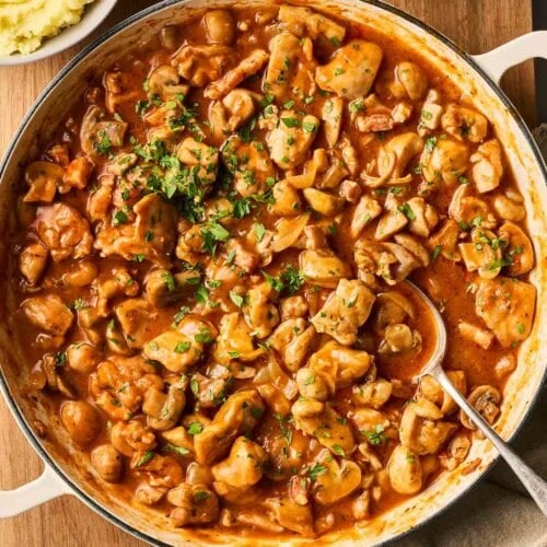 A large pot of French style Chicken Chasseur with chicken and mushrooms, on a table, ready to serve with a spoon, with mashed potatoes to the side.