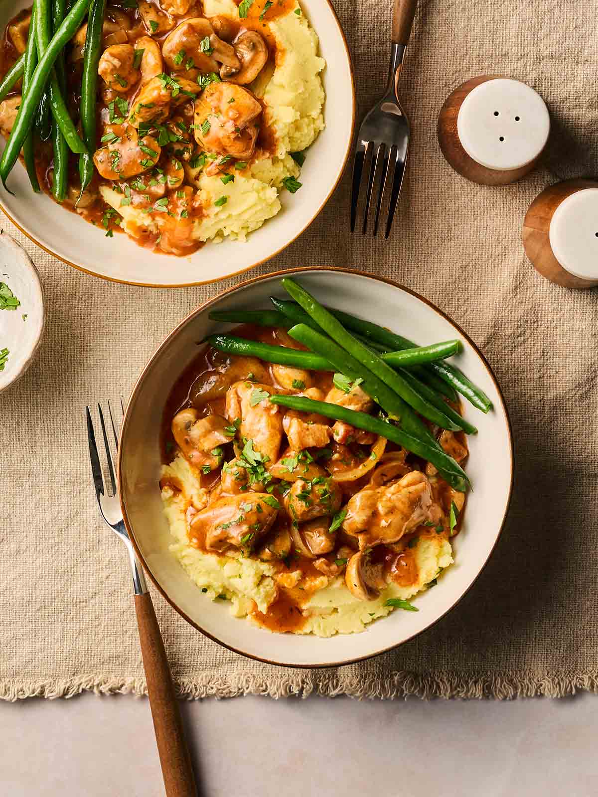 Two bowls of chicken chasseur with mashed potatoes and green beans, on a set table with forks to the side, ready to eat.