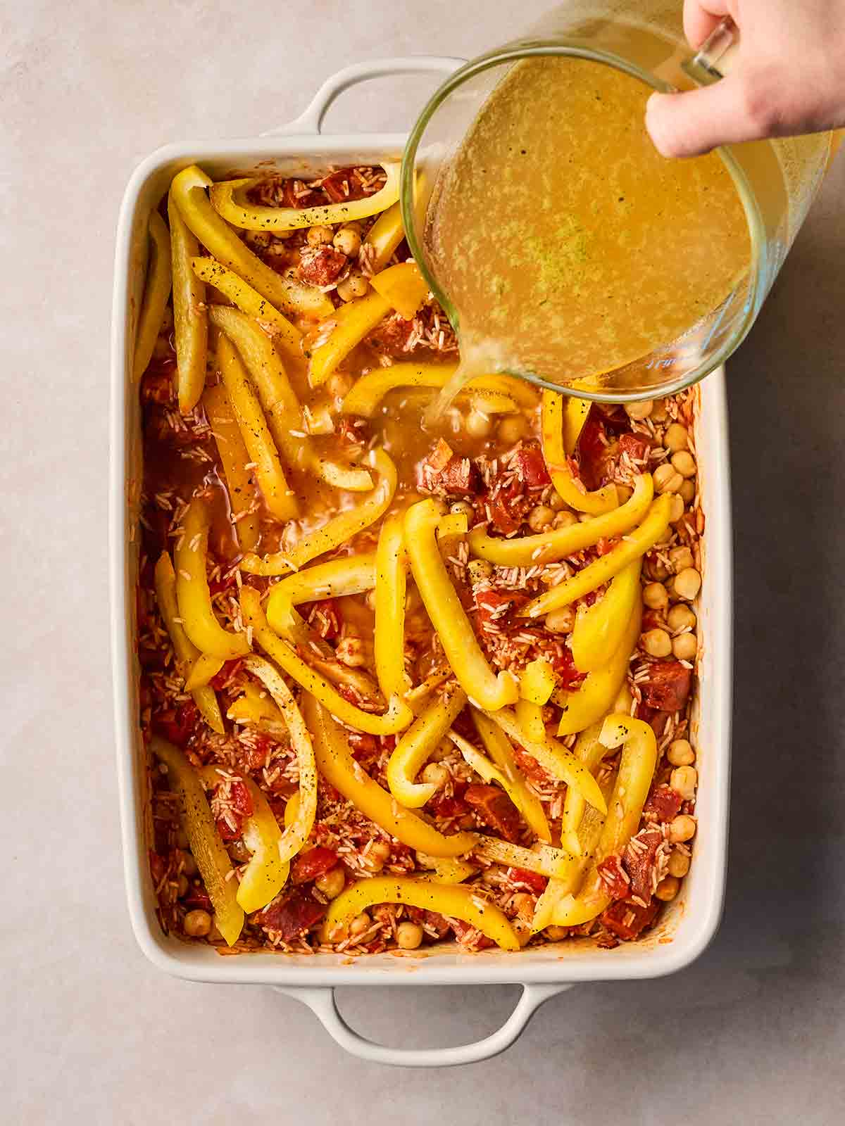 A hand pouring stock from a jug into an oven dish with lot of ingredients, including chorizo, rice, peppers and chickpeas.