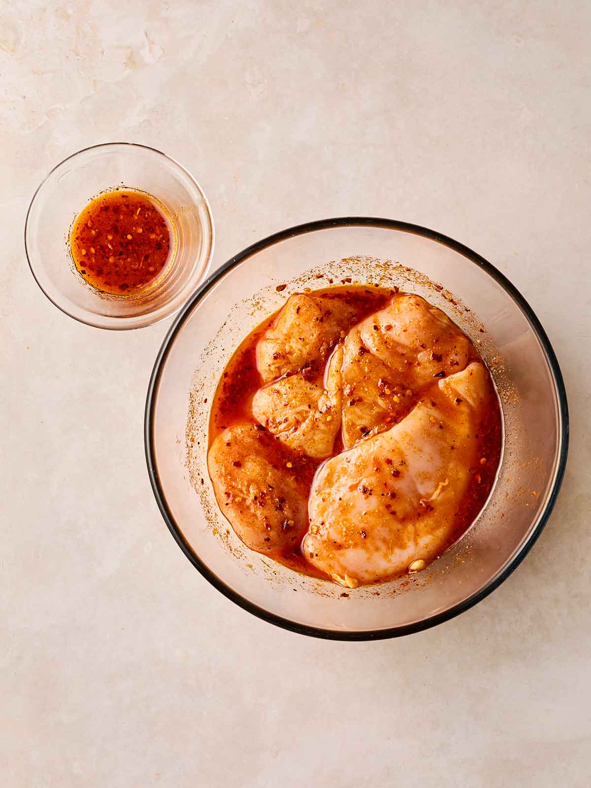 A glass bowl filled with four raw chicken breasts in a red marinade, with a little in a glass to the side, on a white surface.