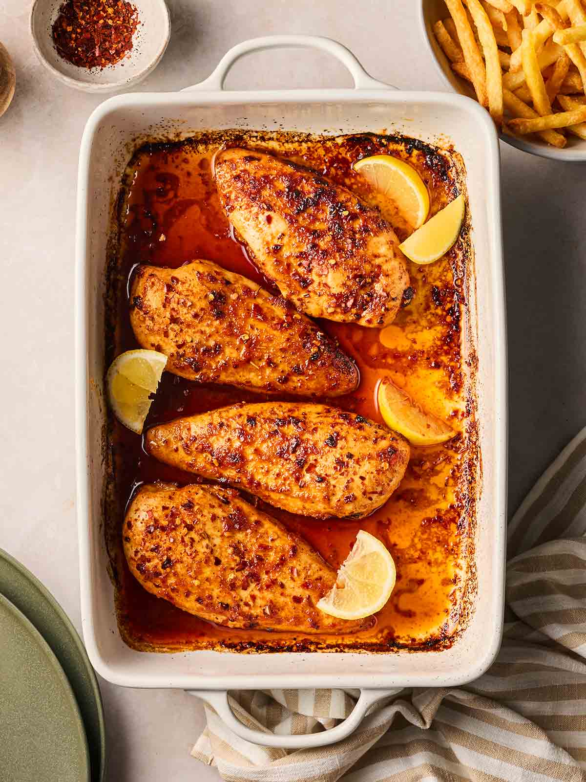 An oven dish with cooked chicken breast in a chilli and honey sauce, known as Divorce Me Chicken, on a table with a bowl of chips to the side.