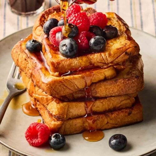 French toast stacked on a plate on a set table, with berries on top and maple syrup being poured over.