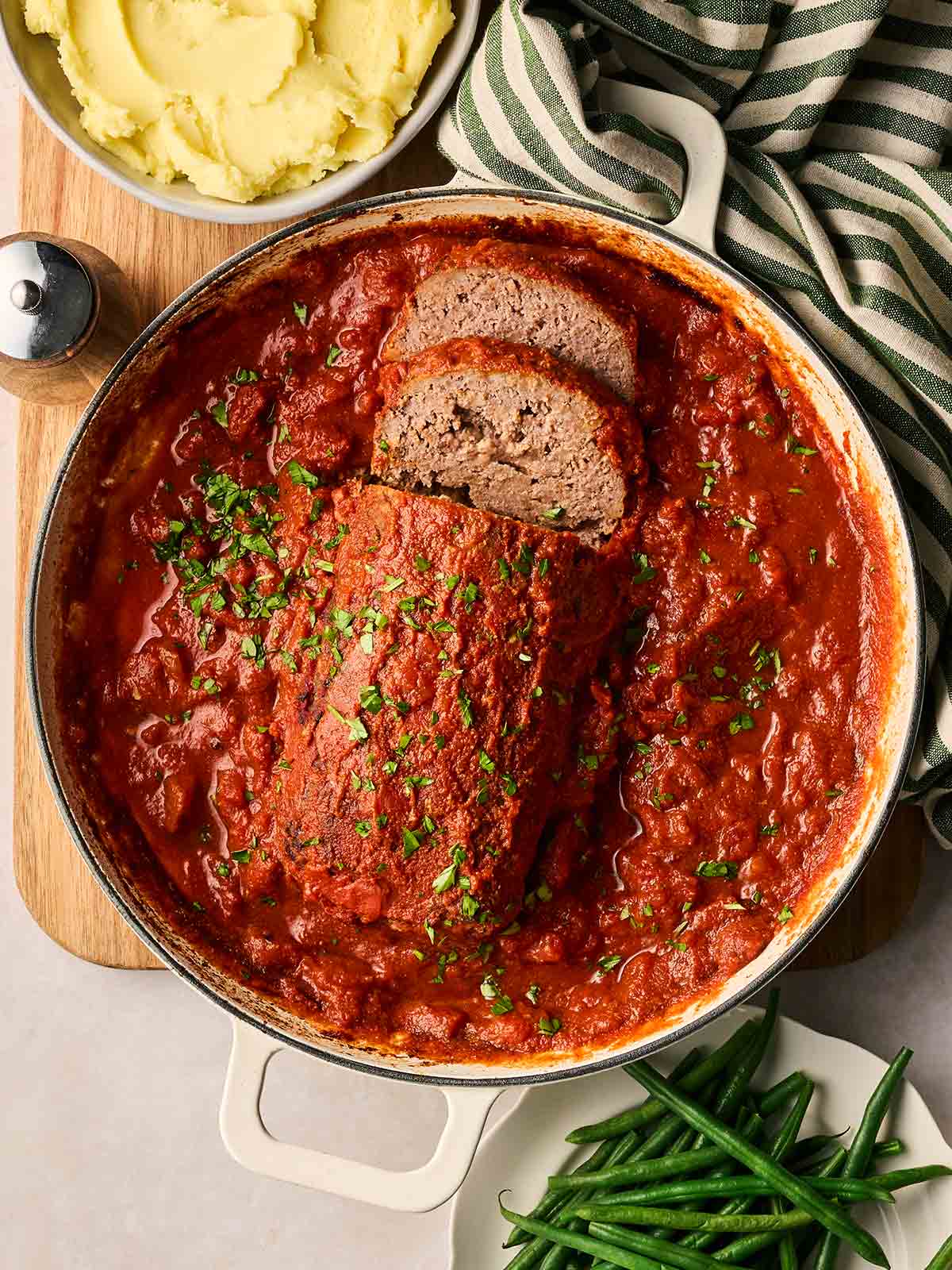 A large pan filled with meatloaf in a rich tomato sauce, on the table with mashed potatoes and green beans on the side, ready to serve.