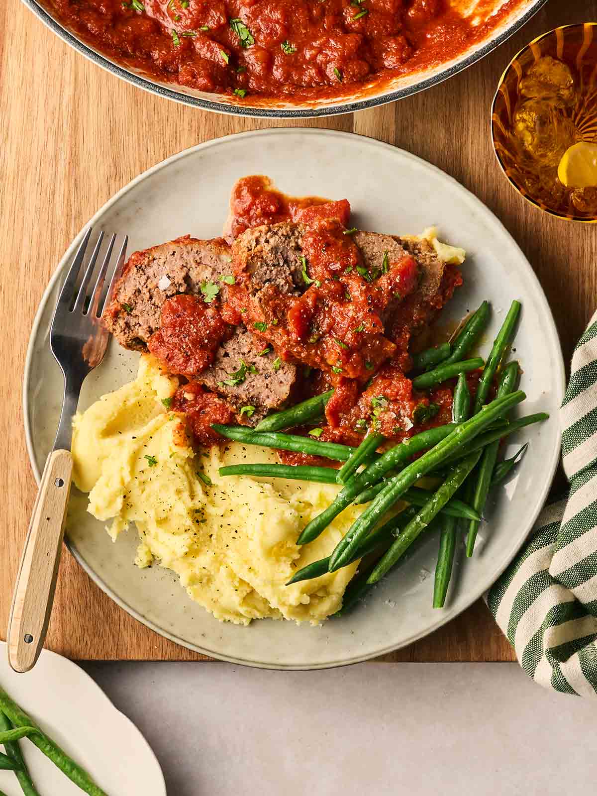 A plate on a table with slices of meatloaf in tomato sauce, greens and mashed potatoes, with a fork ready to serve.