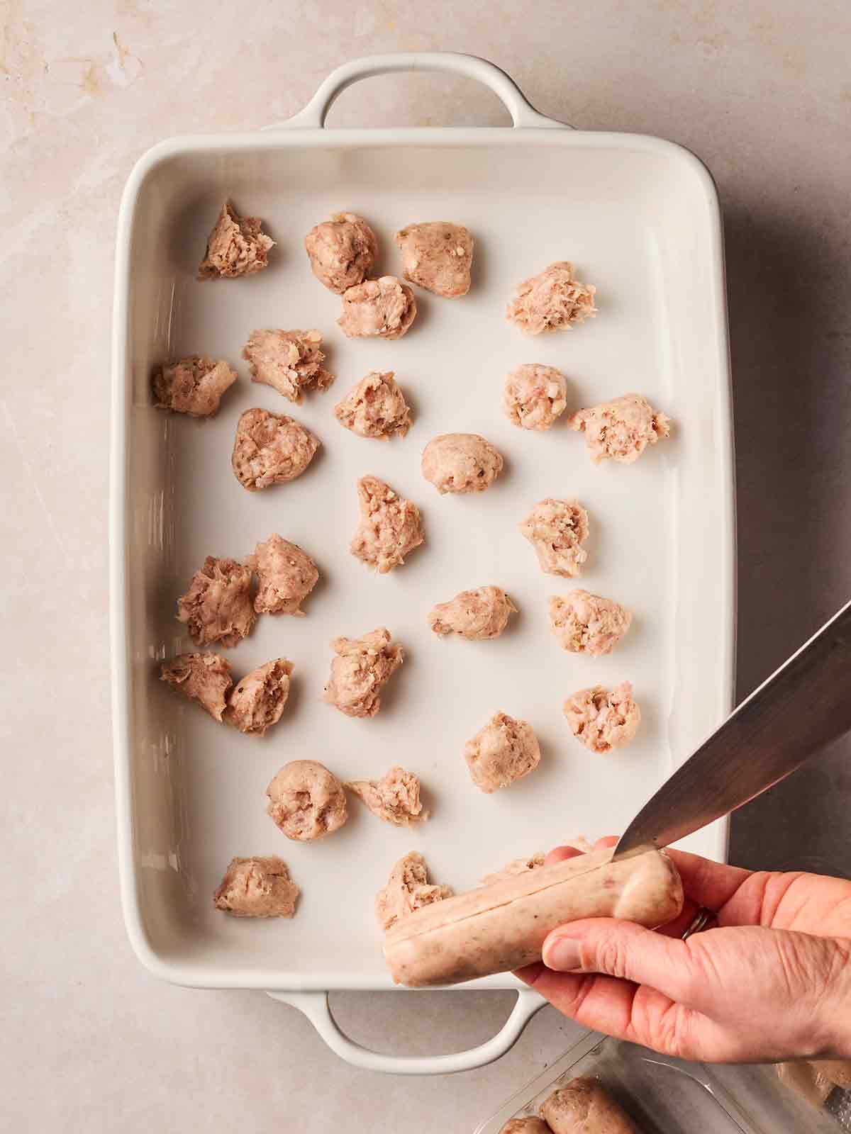 Chunks of raw sausage in an oven dish, with two hands cutting the skin off a sausage with a knife.