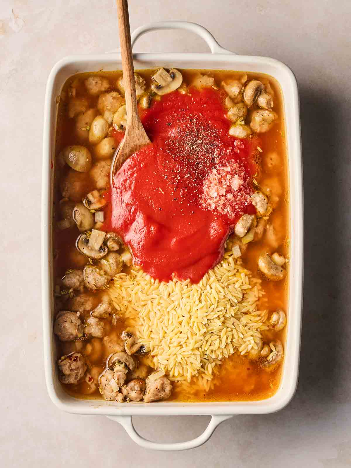 An oven dish with sausage and pasta, with passata and dried orzo over the top, about to be stirred with a wooden spoon.
