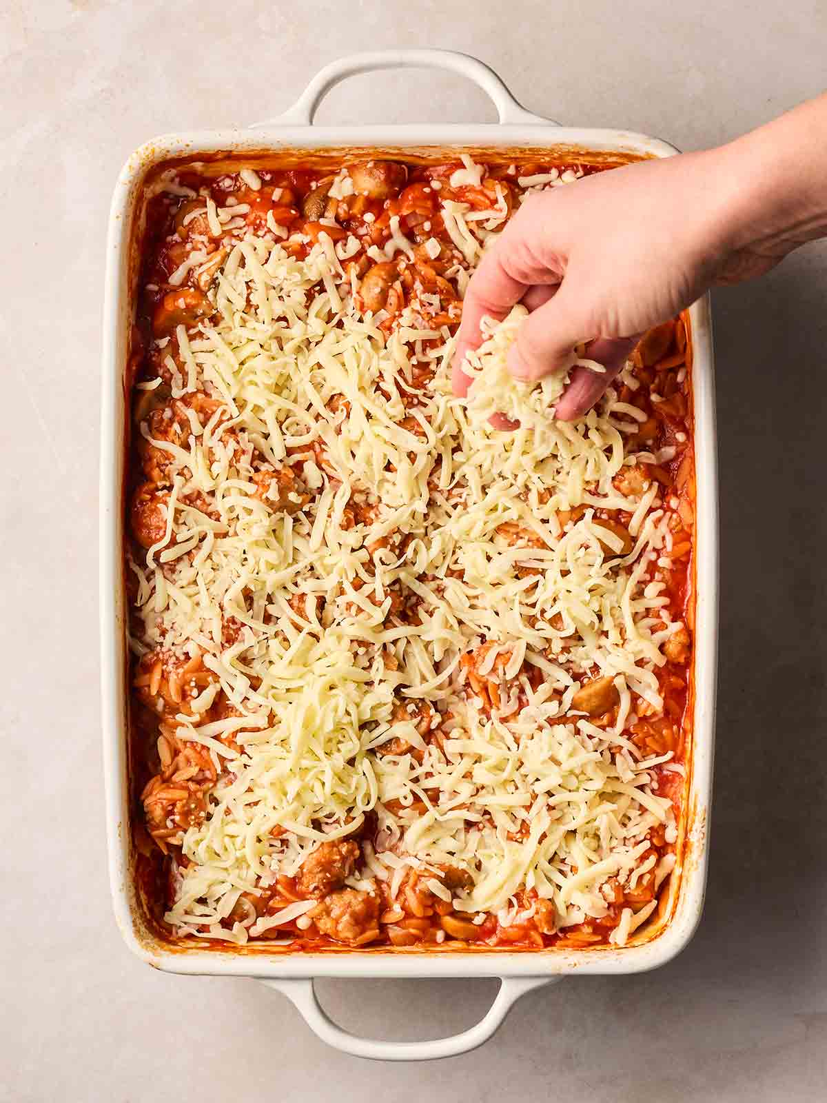 A hand sprinkling grated cheese over a sausage orzo bake dish.