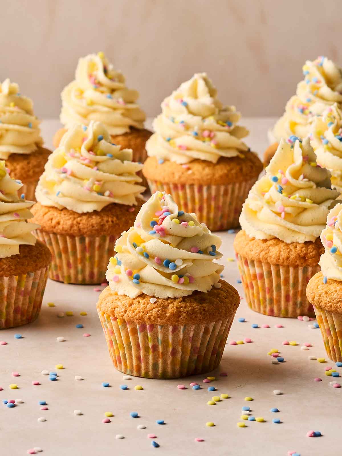Rows of vanilla cupcakes with buttercream icing and sprinkles on top on a white surface.