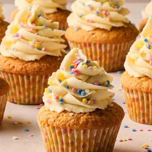 A batch of vanilla cupcakes on a white surface, scattered with sprinkles.