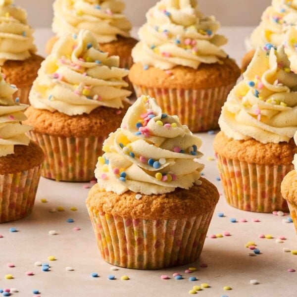 A batch of vanilla cupcakes with piped buttercream frosting on top and colourful sprinkles, on a white surface from the side.