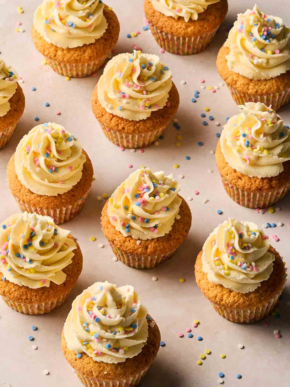 Downwards view of rows of homemade Vanilla Cupcakes , showing cases and the toppings on a white surface.