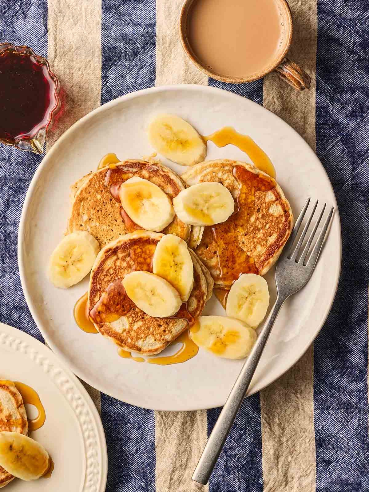 A plate on a blue and white stripy tablecloth with 3 pancakes on with banana and maple syrup, with a fork and a cup of tea to the side.