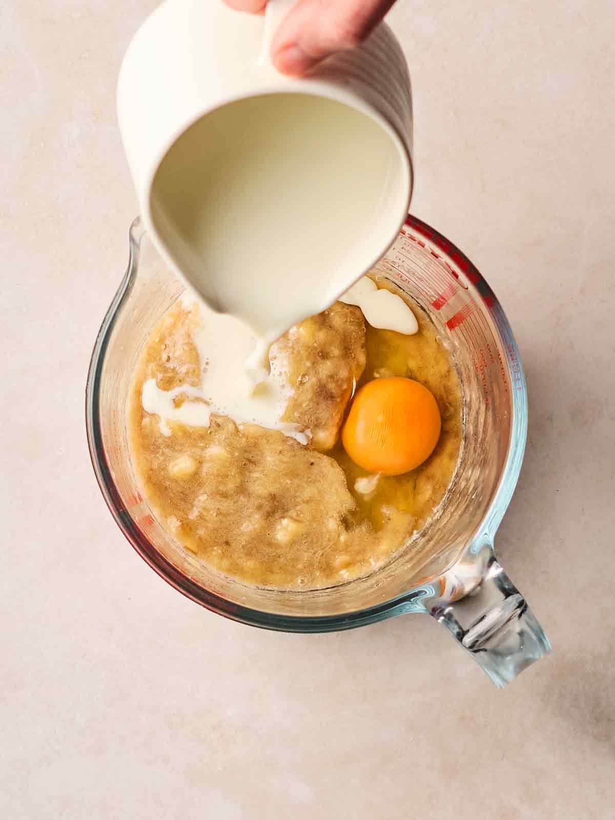 The wet ingredients for making pancakes in a glass jug, with a hand pouring in milk to the mashed banana and eggs.