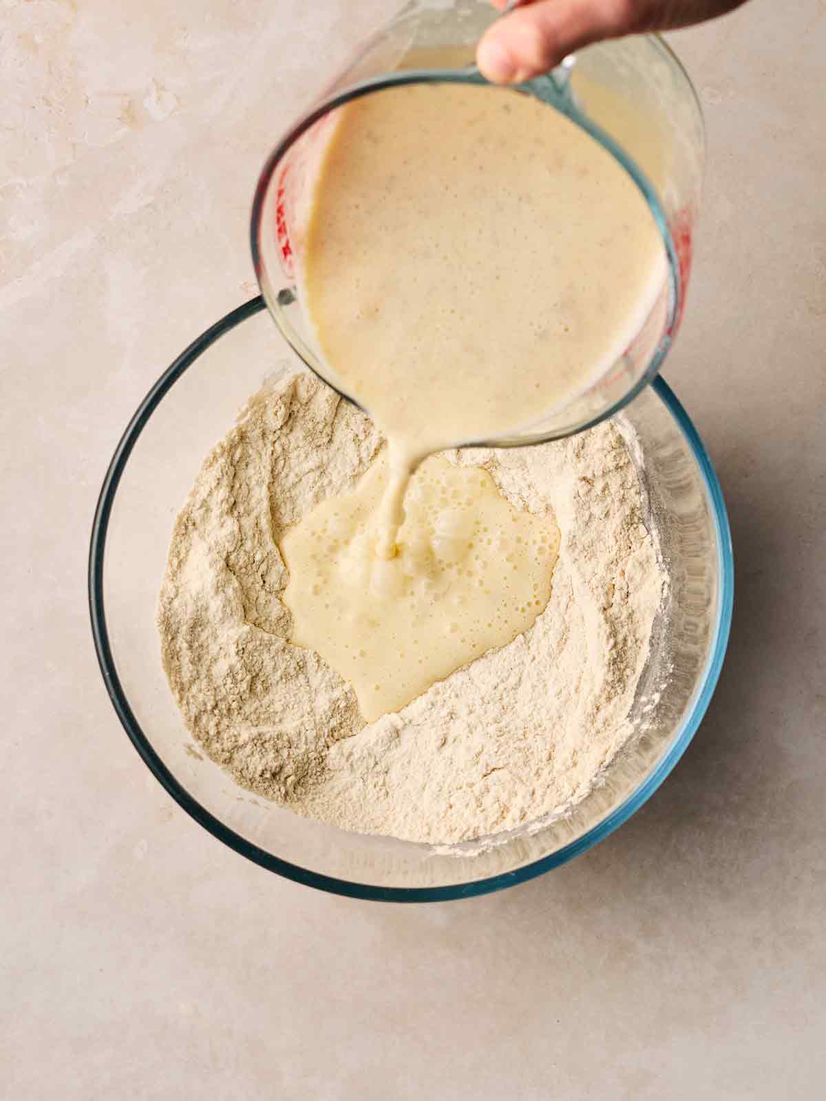 Wet and dry ingredients for making pancakes being poured together in a glass bowl.