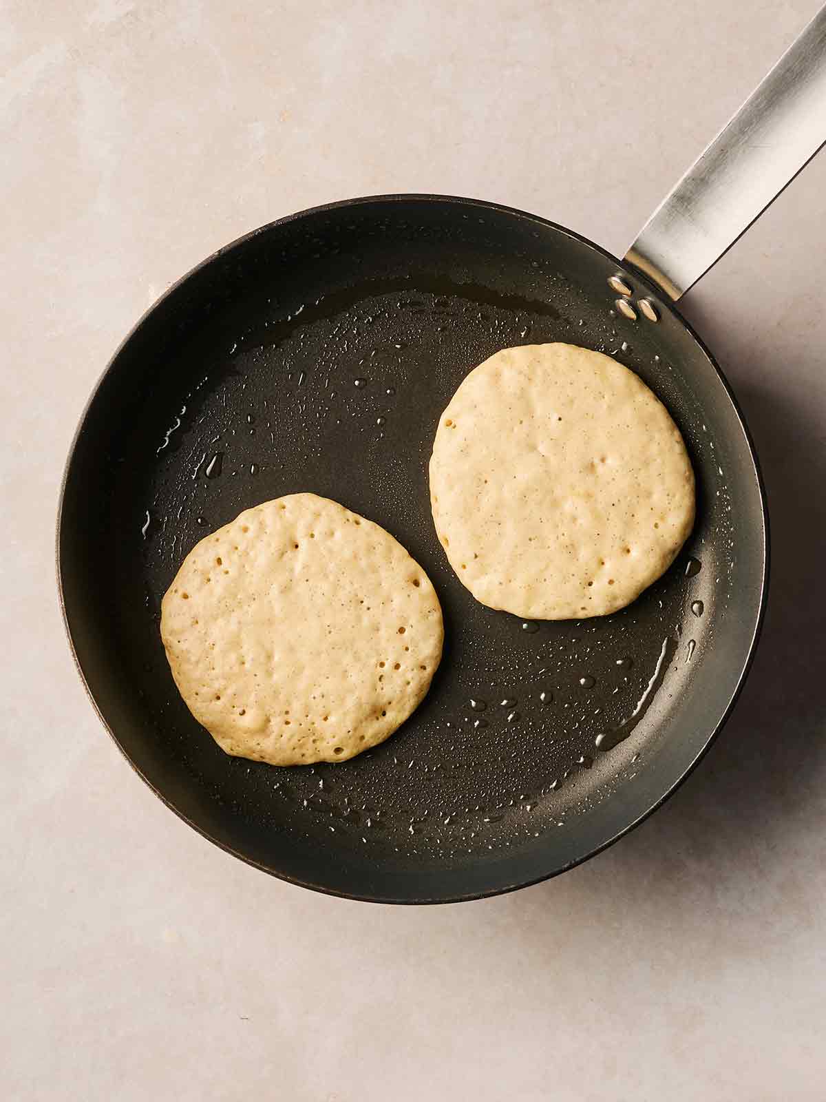A pan on a white surface, with two American-style Banana Pancakes frying in oil, with bubbles coming through the surface.