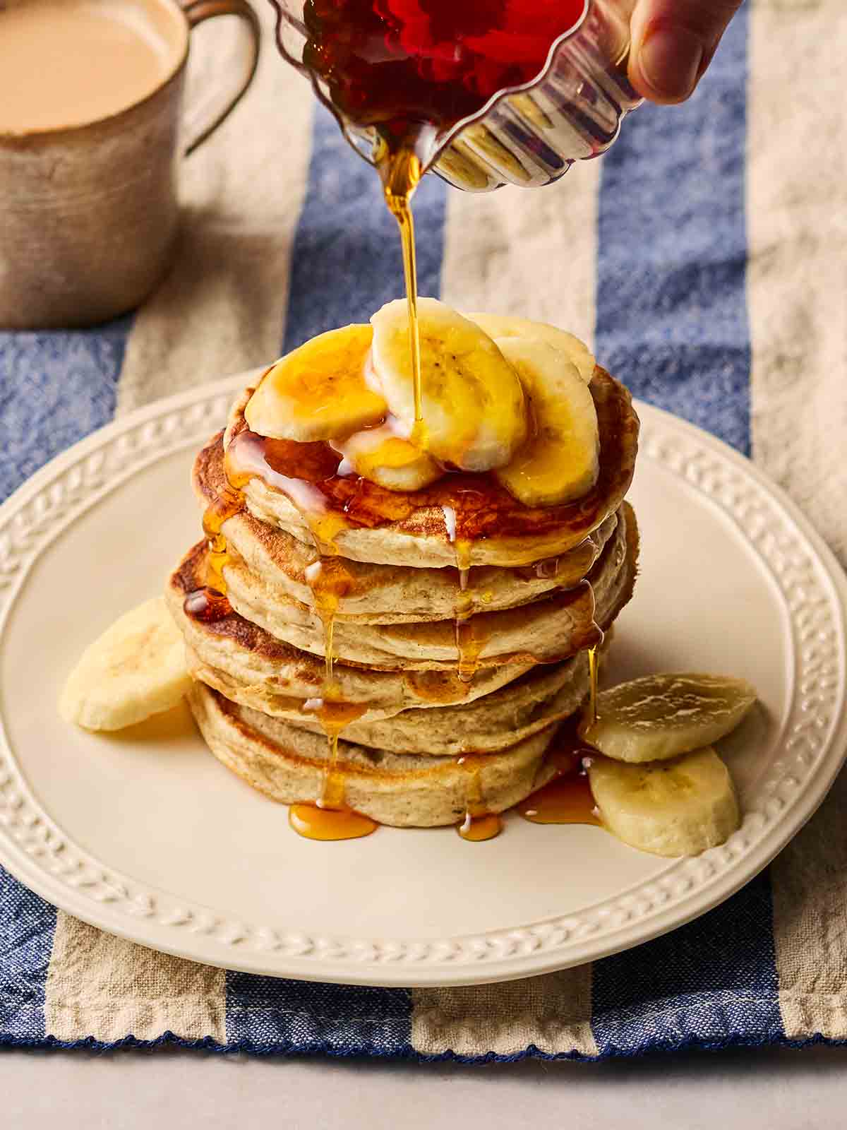 A stack of fluffy Banana Pancakes on a plate, loaded with slices of banana and maple syrup.