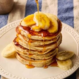 A stack of fluffy American-style Banana Pancakes on a white plate, on a blue and white stripy tablecloth, with slices of banana on top and maple syrup being poured over.