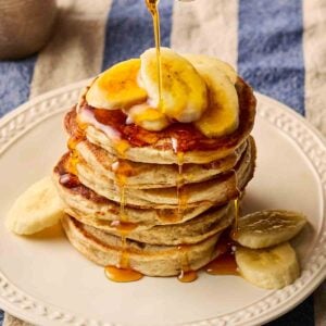 A stack of fluffy American-style Banana Pancakes on a white plate, on a blue and white stripy tablecloth, with slices of banana on top and maple syrup being poured over.