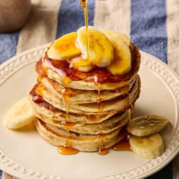 A stack of fluffy American-style Banana Pancakes on a white plate, on a blue and white stripy tablecloth, with slices of banana on top and maple syrup being poured over.