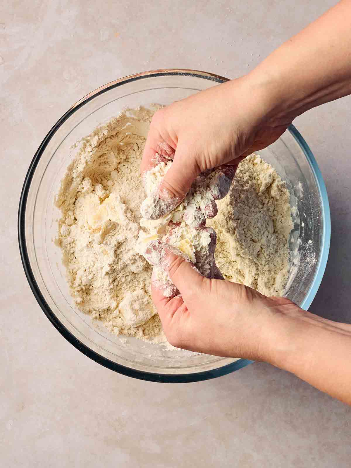 Two hands rubbing flour and butter together over a glass bowl for step 1 in the recipe for Cheese Scones.