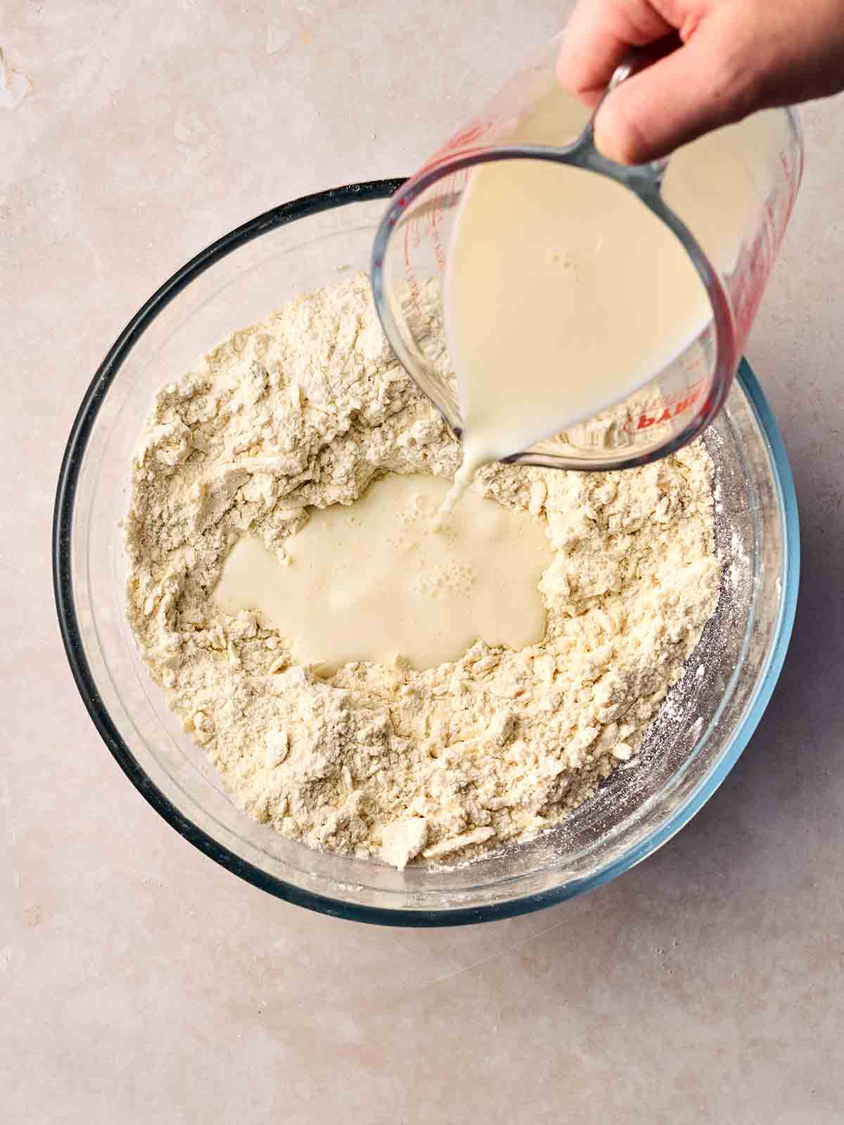 A glass bowl with milk being poured into flour mixture and grated cheese for step 2 in the recipe for Cheese Scones.