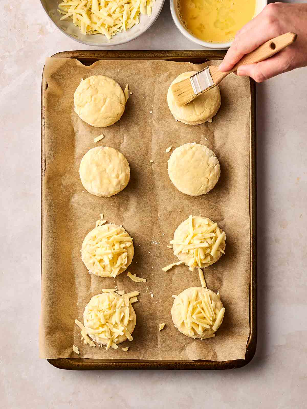 Uncooked Cheese Scones on a baking sheet with a hand brushing on egg.