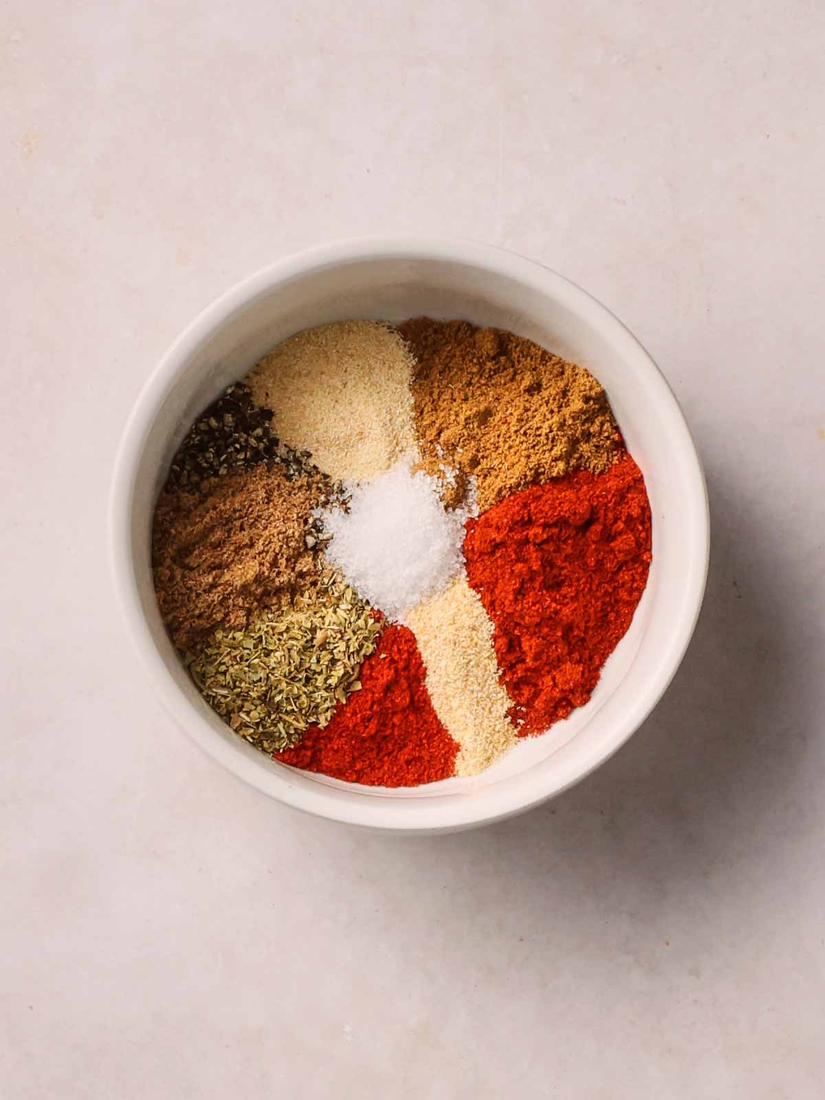 A white bowl on a white surface, with a rainbow of spices inside, ready to be mixed.