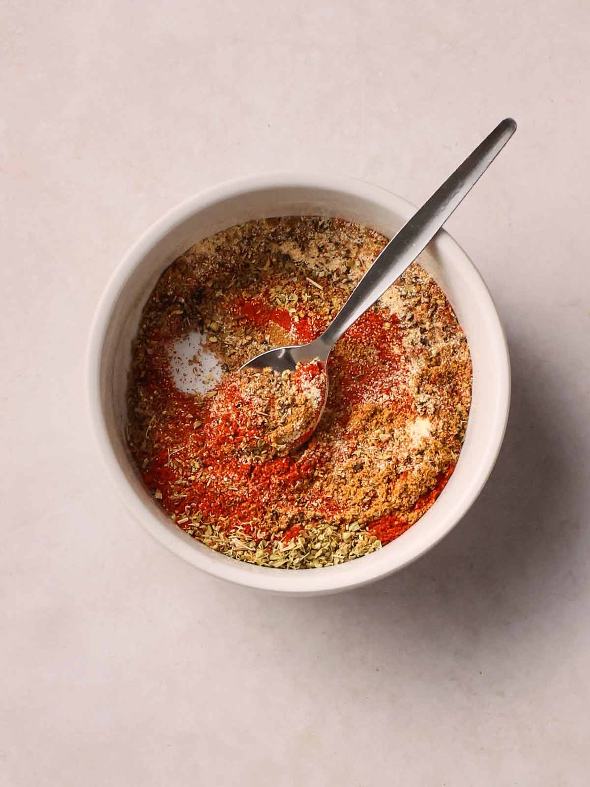 A white bowl on a white surface with a spoon inside, with a variety of spices mixed together.
